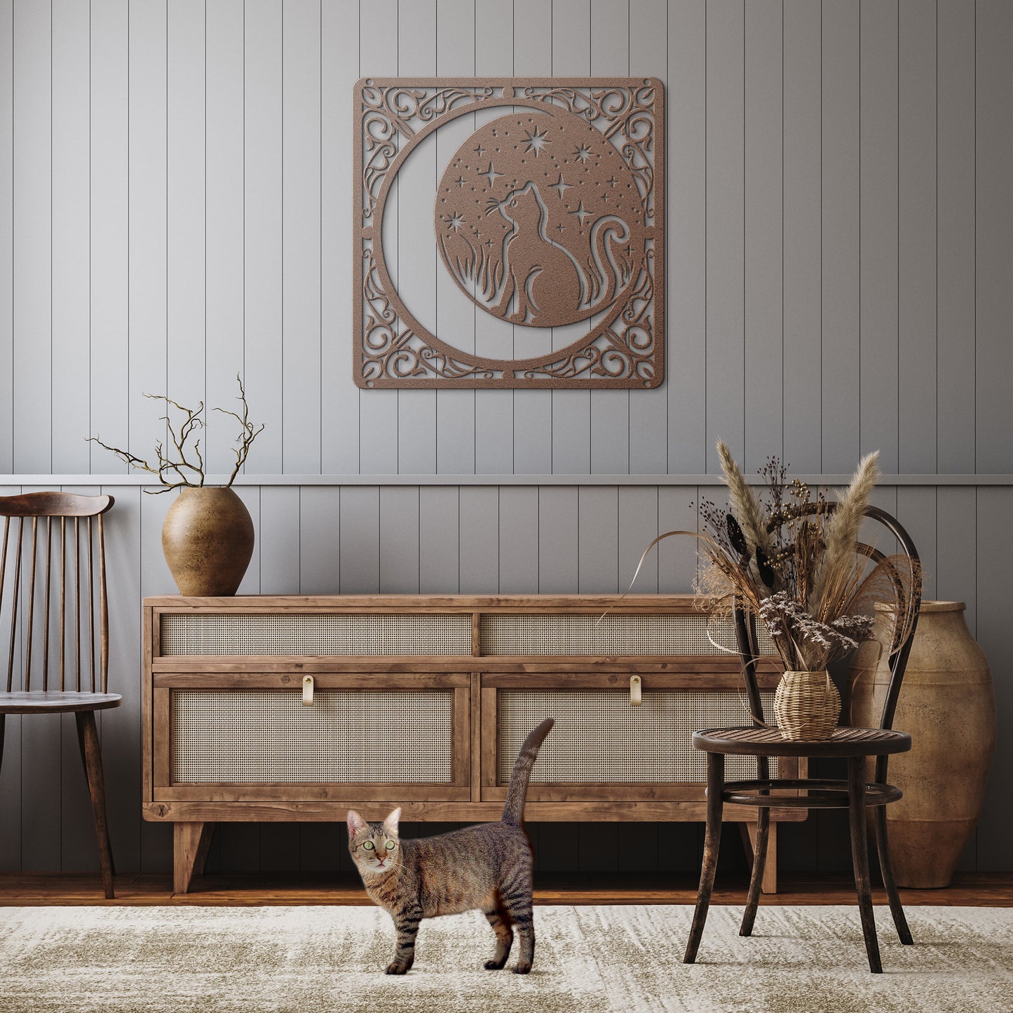 A copper home decor metal sign of a cat inside a crescent moon next to a field of stars. All of this is bordered by floral vines. It hangs on the wall of a rustic hallway above a tabby cat.