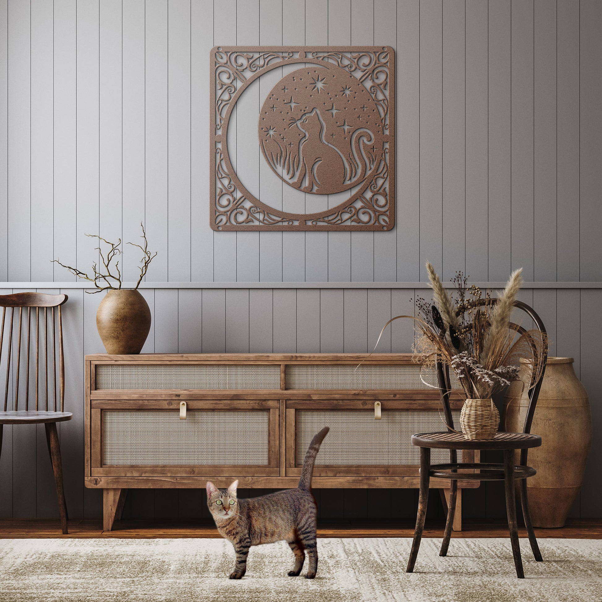 A copper home decor metal sign of a cat inside a crescent moon next to a field of stars. All of this is bordered by floral vines. It hangs on the wall of a rustic hallway above a tabby cat.
