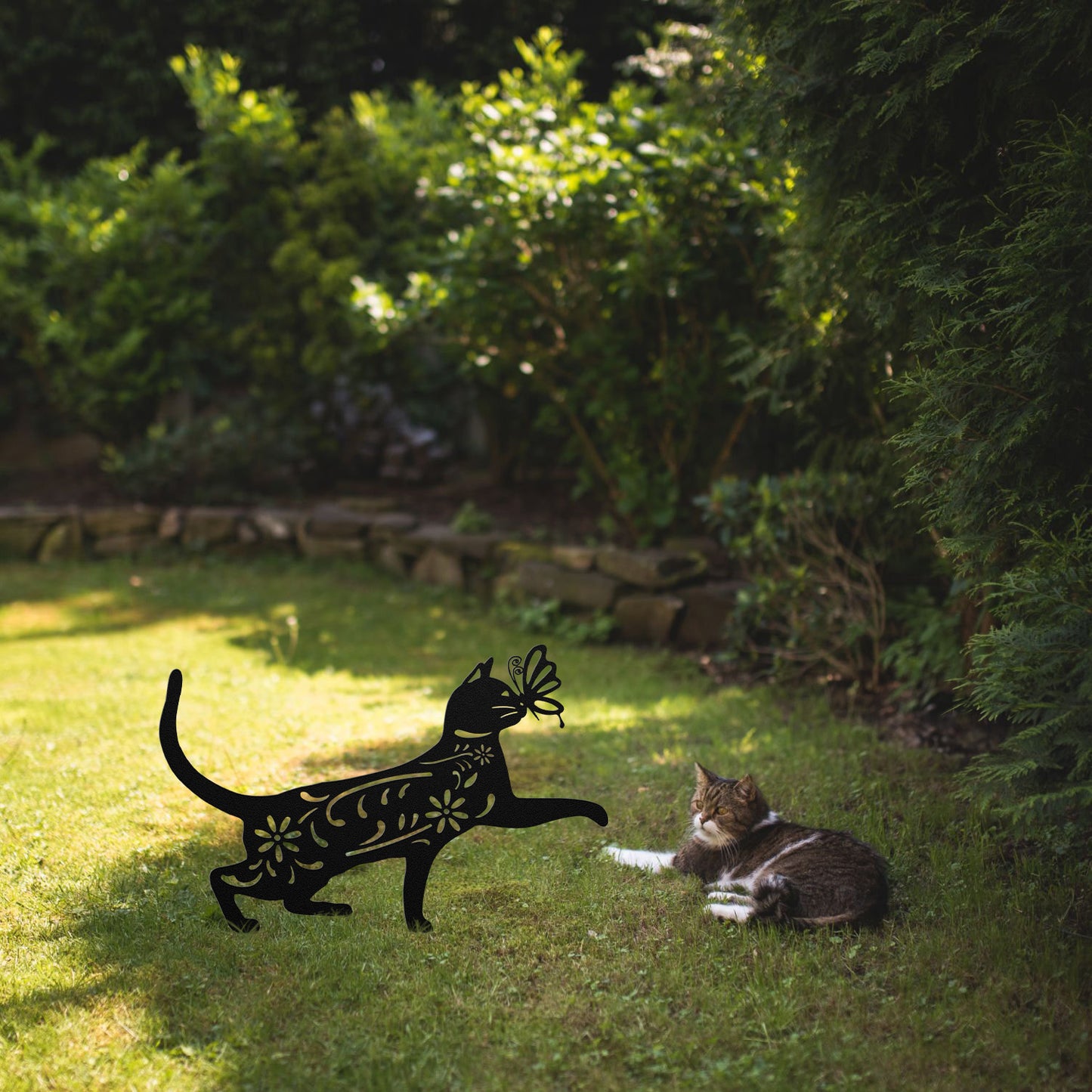 A black yard/garden stake of a cat with a butterfly on it nose and floral elements on it body. It is placed on a grass lawn near a tabby cat.