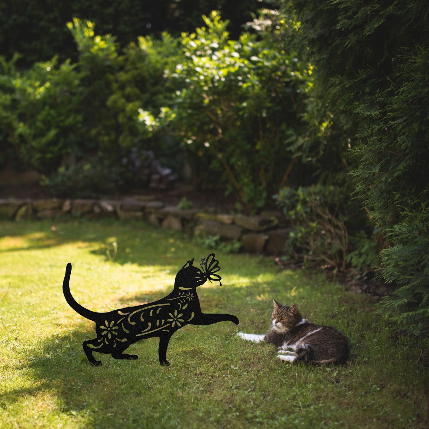 A black yard/garden stake of a cat with a butterfly on it nose and floral elements on it body. It is placed on a grass lawn near a tabby cat.