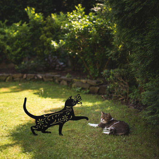 A black yard/garden stake of a cat with a butterfly on it nose and floral elements on it body. It is placed on a grass lawn near a tabby cat.