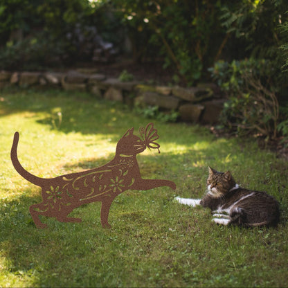 A copper yard/garden stake of a cat with a butterfly on it nose and floral elements on it body. It is placed on a grass lawn near a tabby cat.