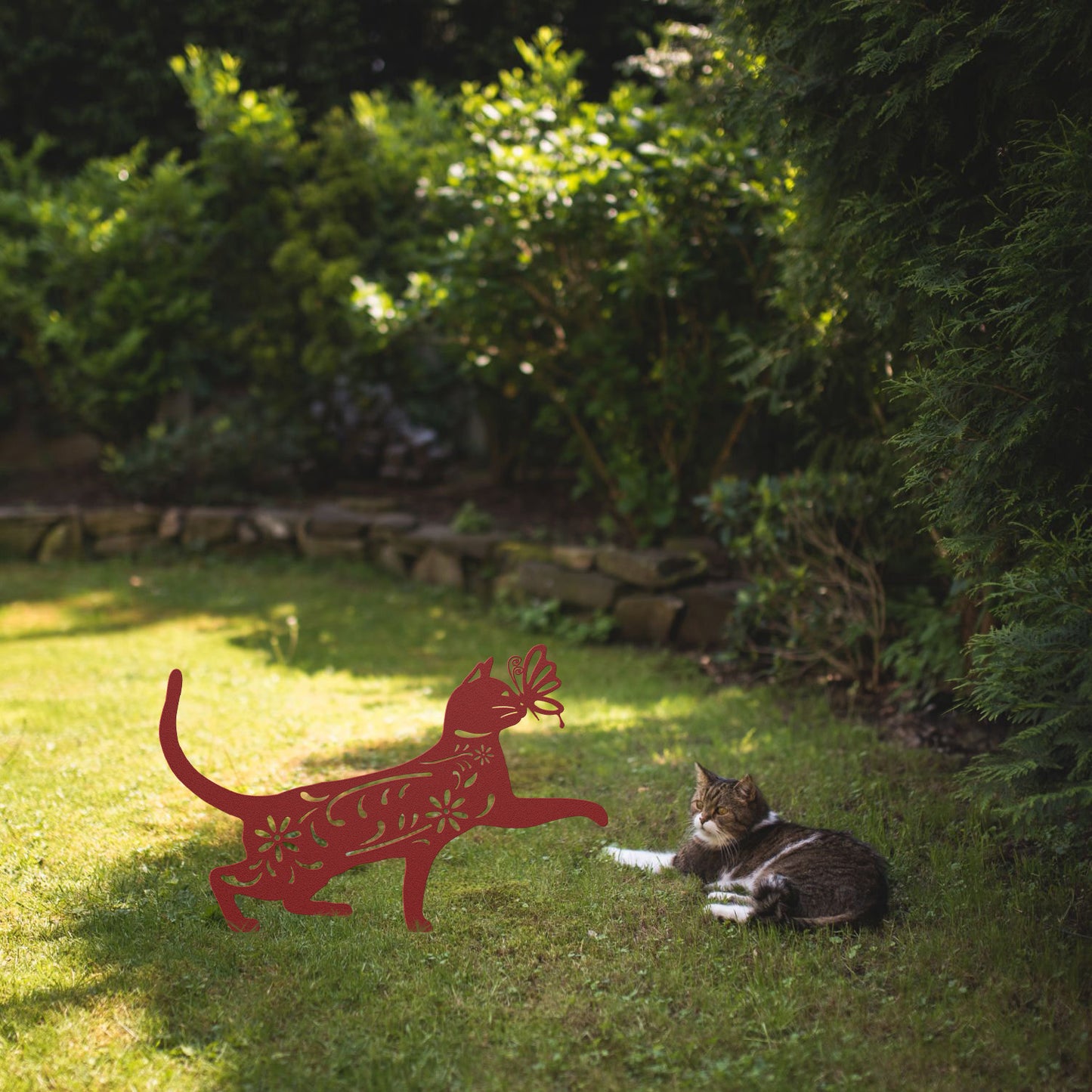 A red yard/garden stake of a cat with a butterfly on it nose and floral elements on it body. It is placed on a grass lawn near a tabby cat.