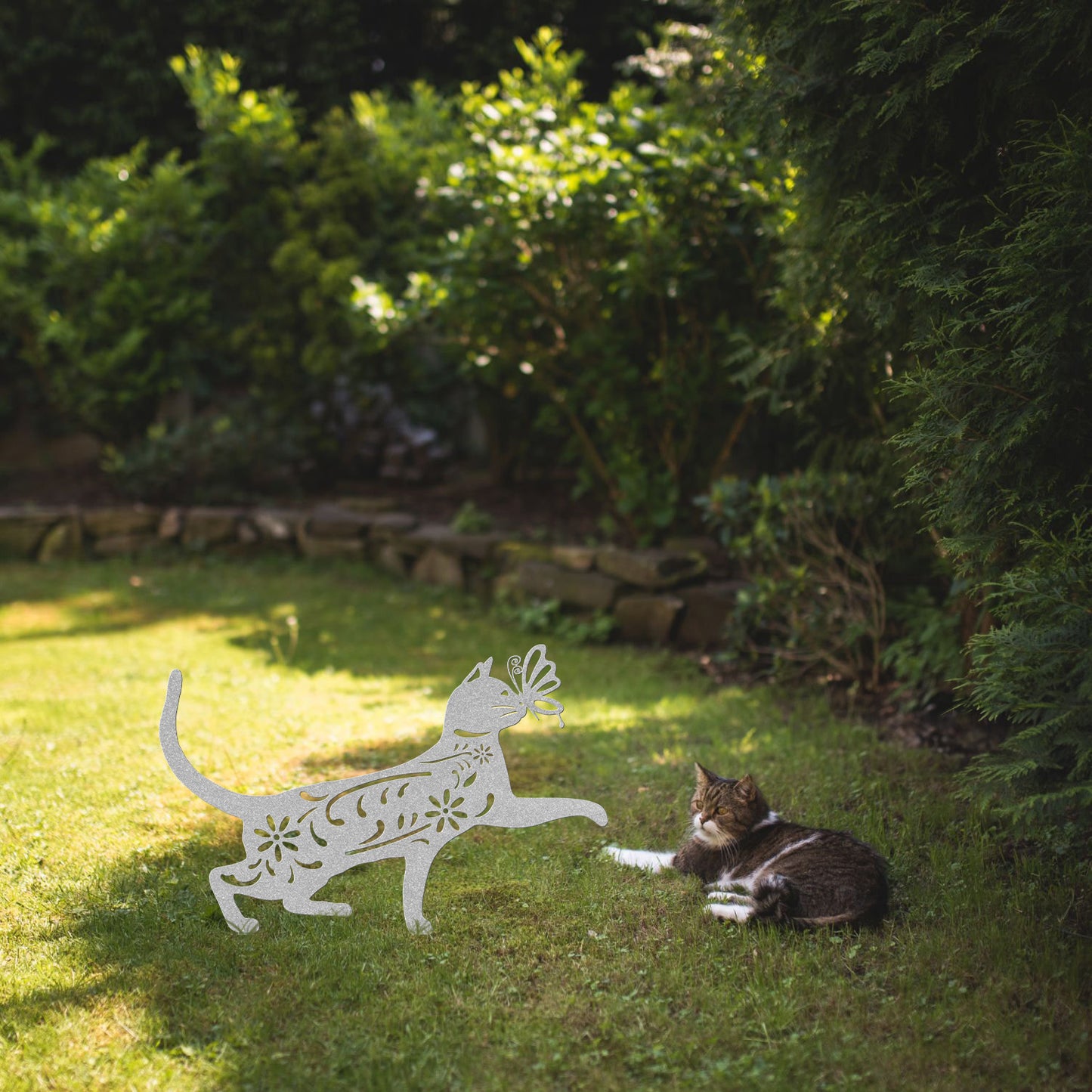 A silver yard/garden stake of a cat with a butterfly on it nose and floral elements on it body. It is placed on a grass lawn near a tabby cat.