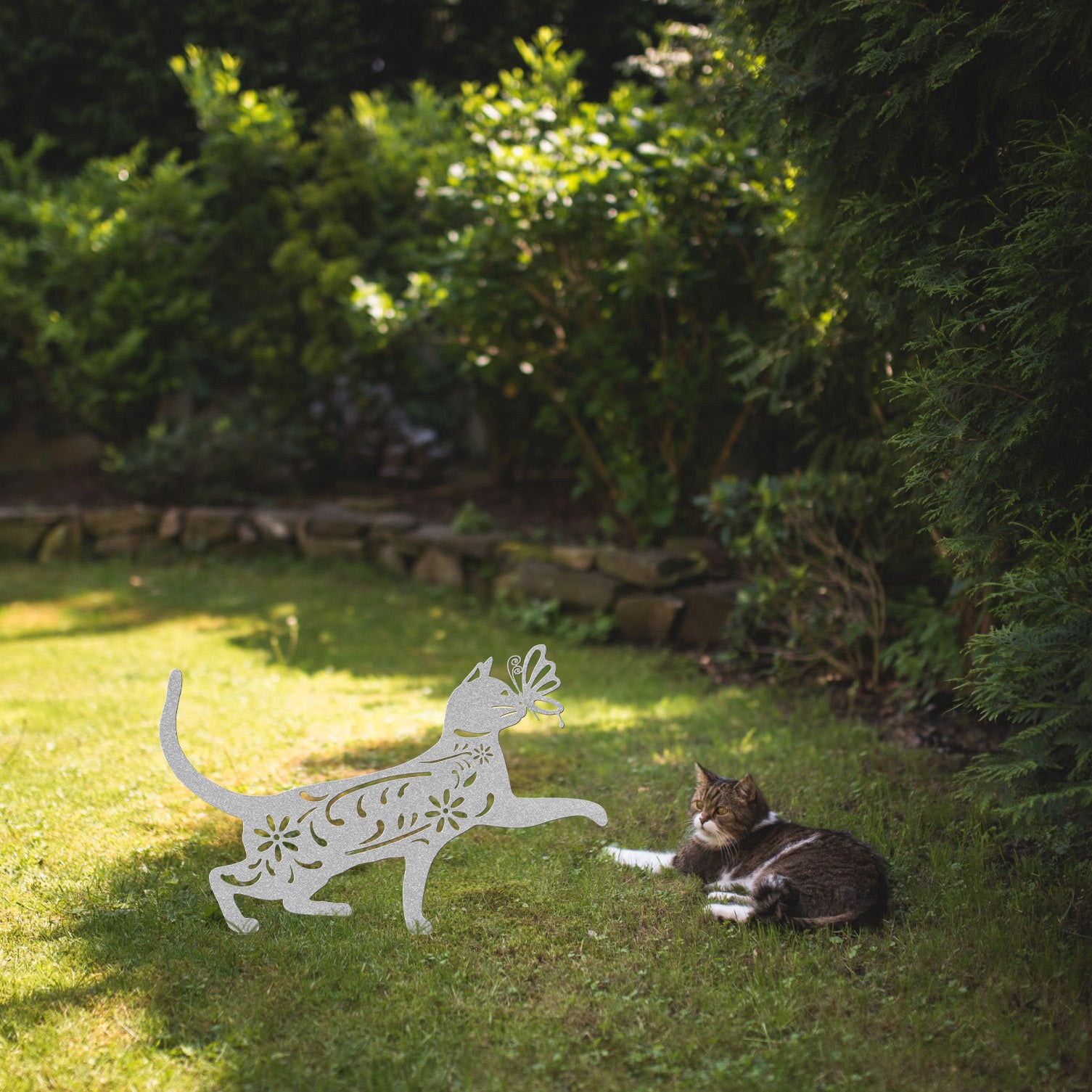 A silver yard/garden stake of a cat with a butterfly on it nose and floral elements on it body. It is placed on a grass lawn near a tabby cat.