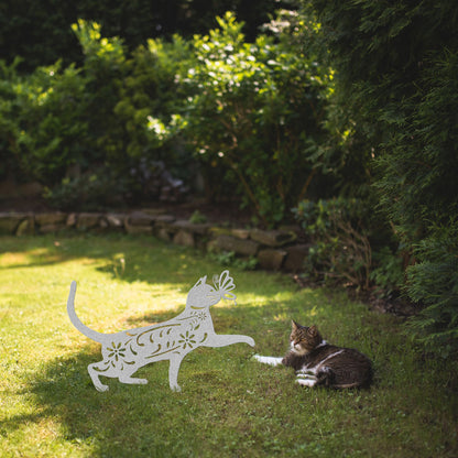 A silver yard/garden stake of a cat with a butterfly on it nose and floral elements on it body. It is placed on a grass lawn near a tabby cat.