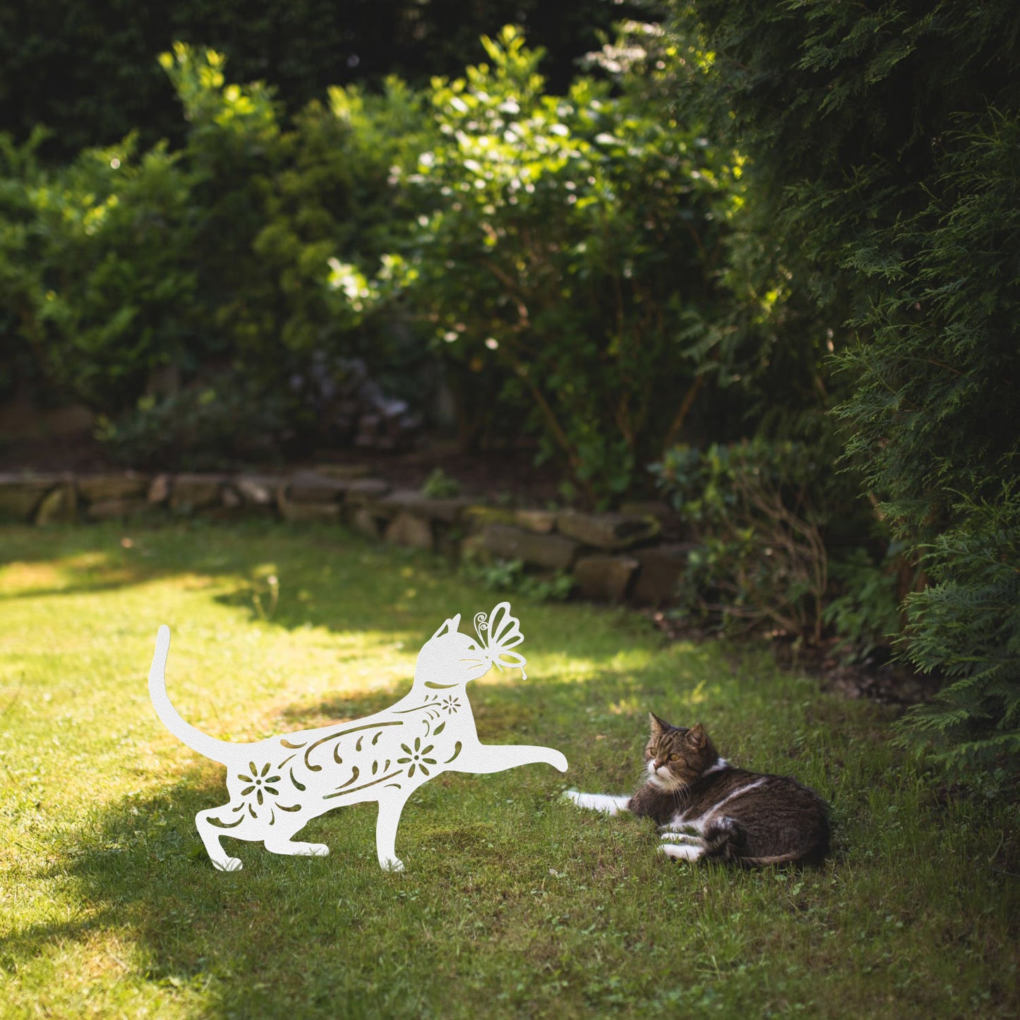 A white yard/garden stake of a cat with a butterfly on it nose and floral elements on it body. It is placed on a grass lawn near a tabby cat.