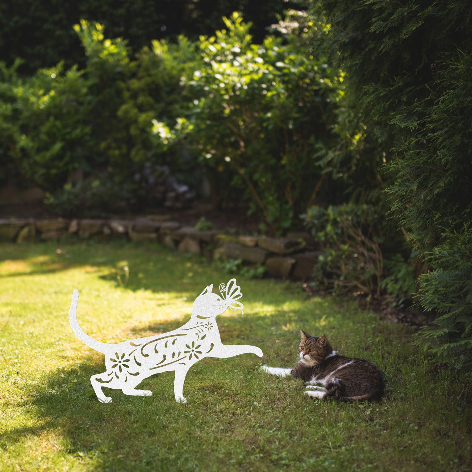 A white yard/garden stake of a cat with a butterfly on it nose and floral elements on it body. It is placed on a grass lawn near a tabby cat.