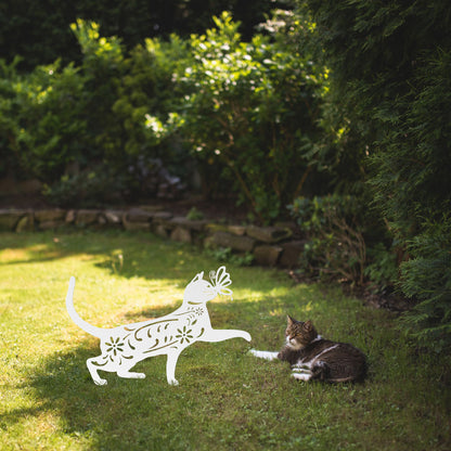 A white yard/garden stake of a cat with a butterfly on it nose and floral elements on it body. It is placed on a grass lawn near a tabby cat.